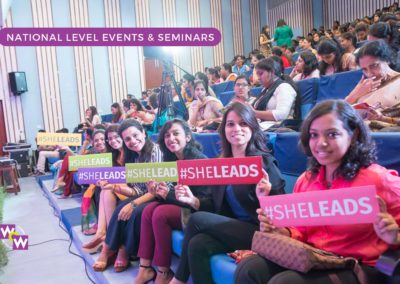 A group of young women seated in an auditorium during a national WOW seminar and smiling holding placards that read She Leads